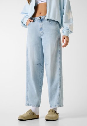 Osoba w jasnoniebieskich szerokich dżinsach, krótkim topie, jasnoniebieskiej bluzie z białymi gwiazdkami, białych skarpetkach i jasnobrązowych butach wsuwanych.