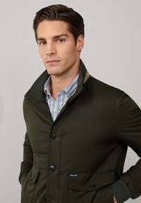 Uomo con capelli castano scuro che indossa una giacca verde oliva sopra una camicia a righe blu e bianca con colletto, in piedi davanti a uno sfondo grigio uniforme.