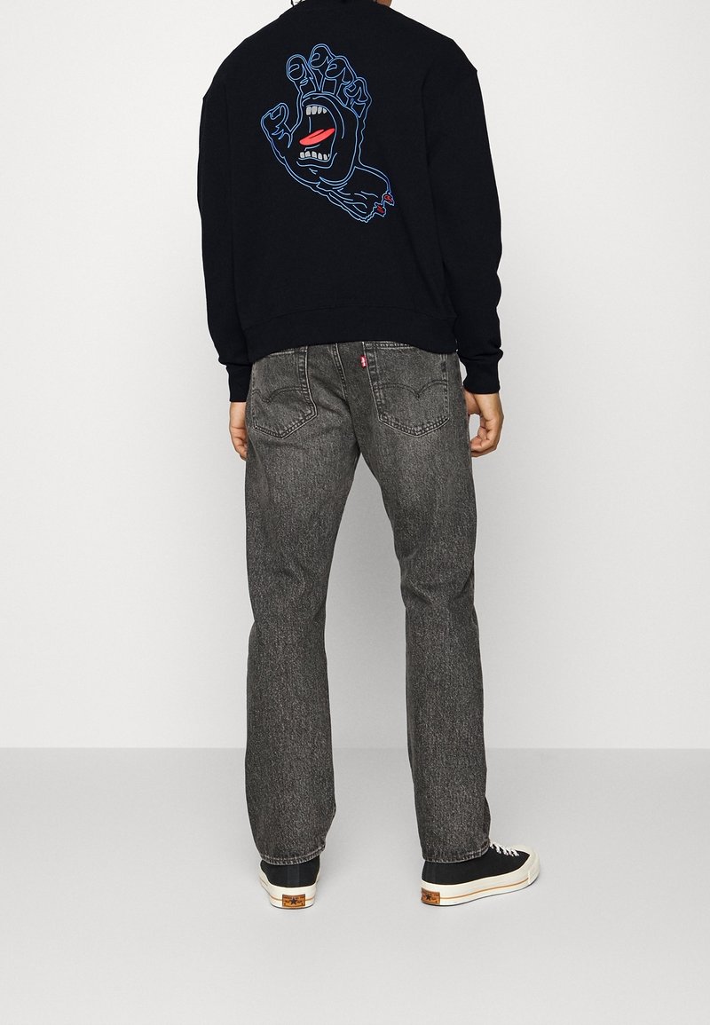Personne portant un sweat-shirt noir avec un graphique d'une main bleue à bouche ouverte dans le dos, un jean gris et des baskets noires à semelles blanches.