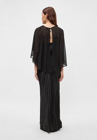 Dlouhé černé šaty s průsvitným černým pláštíkem. Má hladkou saténovou texturu a detail s výstřihem na zádech. Přiléhavý střih.