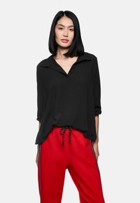 Schwarzes Langarmshirt mit Kragen und Knöpfen, kombiniert mit roten Hosen mit Seitestreifen und einem Kordelzugbund.