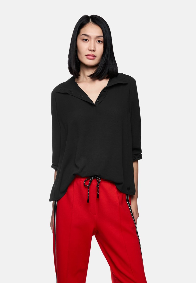 Schwarzes Langarmshirt mit Kragen und Knöpfen, kombiniert mit roten Hosen mit Seitestreifen und einem Kordelzugbund.
