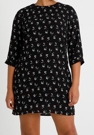 Femme portant une robe noire avec de petits motifs floraux blancs et des manches trois-quarts, debout devant un fond uni.