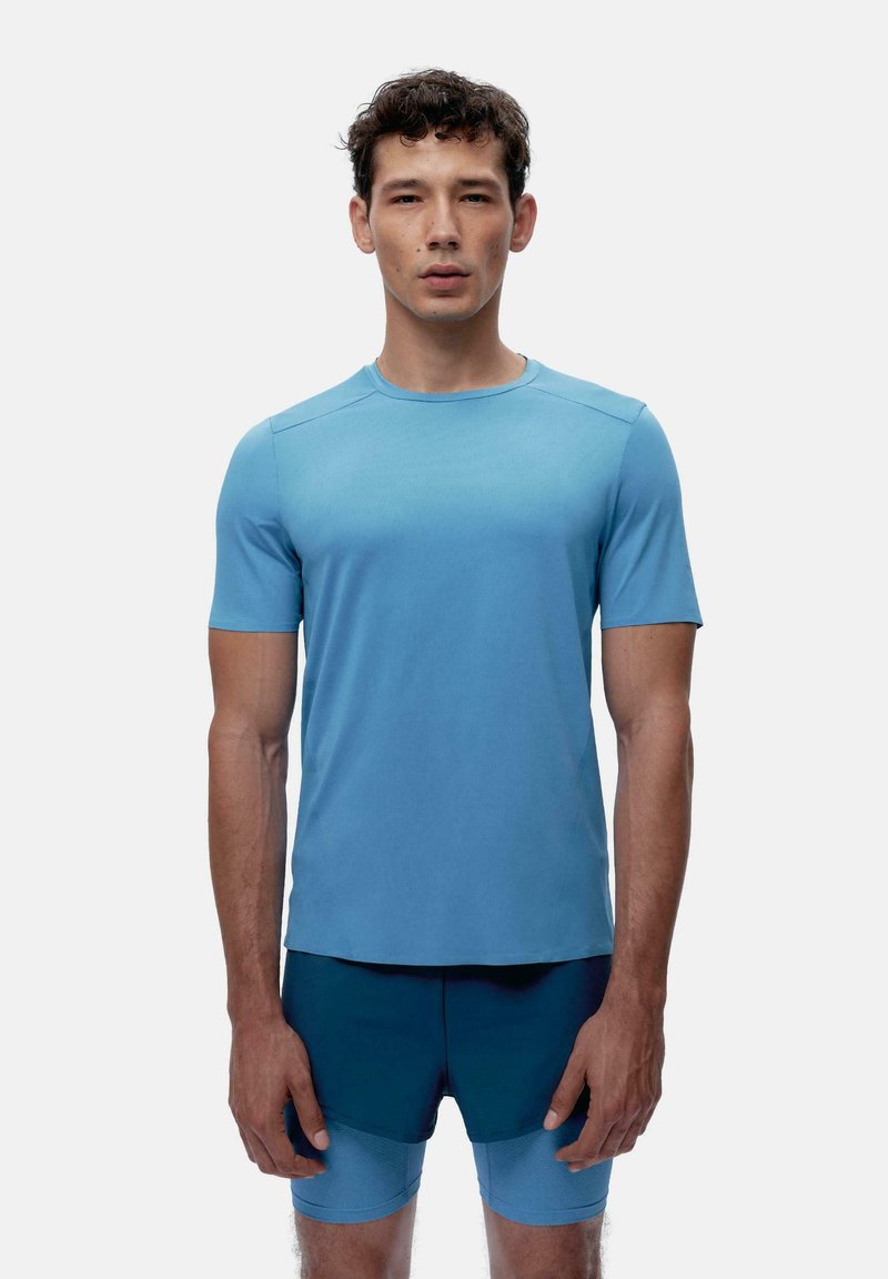 Šviesiai mėlyna trumpomis rankovėmis sportinė marškinėliai su apvaliu kaklu, pagaminti iš lygaus audinio. Pavaizduoti su tamsiai mėlynomis sportinėmis šortais, minimalizmo dizainu.