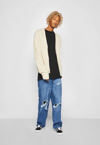 Cardigan en tricot beige avec deux boutons, porté sur un t-shirt noir, associé à un jean bleu déchiré et des chaussures en toile noires.