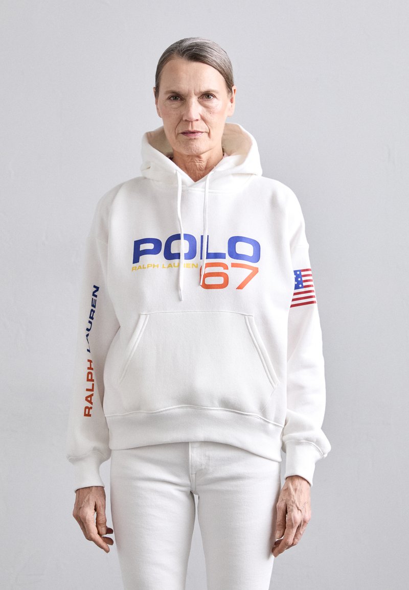 Balta kapuce ar uzrakstu "POLO 67" zilā un oranžā krāsā. Piedāvā "RALPH LAUREN" uzrakstu zilā krāsā uz piedurknes un nelielu Amerikas karoga akcentu.