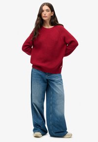 Maglione rosso lavorato a maglia con scollo rotondo e orlo a coste, abbinato a jeans in denim blu a gamba larga. Vestibilità casual, tessuto testurizzato e silhouette rilassata.