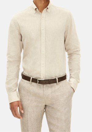 Uomo con camicia beige a maniche lunghe abbottonata infilata in pantaloni beige coordinati con cintura marrone scuro, mani rilassate ai lati.