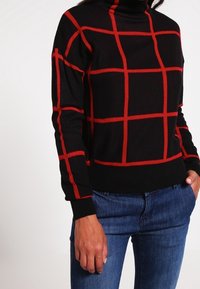 Schwarzer Rollkragenpullover mit rotem Karomuster, getragen mit blauen Jeans, die Hand lässig in der Tasche.