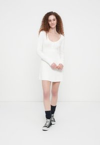 Λευκό φόρεμα πουλόβερ με μακριά μανίκια, ριμπ υφή, στρογγυλή λαιμόκοψη και εφαρμοστή σιλουέτα, συνδυασμένο με μαύρα παπούτσια τύπου κοντά και πλεκτές κάλτσες.