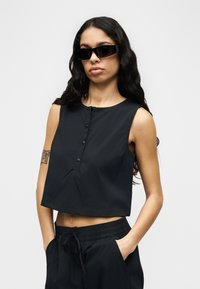 Νεαρή γυναίκα με μακριά κυματιστά μαλλιά φοράει μαύρο αμάνικο τοπ crop, αντίστοιχο παντελόνι και ορθογώνια μαύρα γυαλιά ηλίου, στέκεται με τα χέρια στις τσέπες.