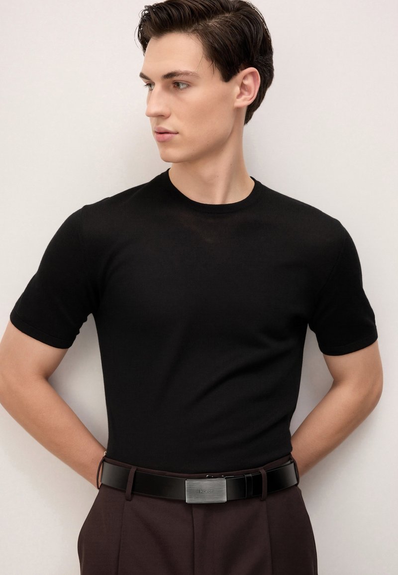 Junger Mann mit kurzen dunklen Haaren, der ein schwarzes kurzärmeliges Hemd und dunkle Hosen mit einem schwarzen Gürtel trägt und nach links schaut.