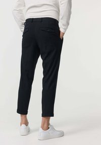 Pantalon noir fuselé avec poches latérales et arrière, ourlets retroussés, associé à des baskets blanches. Le tissu semble lisse avec une coupe structurée.