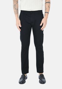 Pantaloni neri slim-fit con una texture liscia, dotati di chiusura con bottone e tasche frontali. Indossati con mocassini neri.