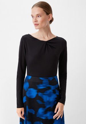 Schwarzes Langarmoberteil mit V-Ausschnitt und einem leichten Drehdetail in der Mitte, kombiniert mit einem blauen gemusterten Rock mit abstrakten Formen.