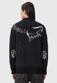 Zwarte wollen cardigan met witte spinnenweb- en hartmotieven, afgewerkt met een textuur en een sjaalkraagontwerp.