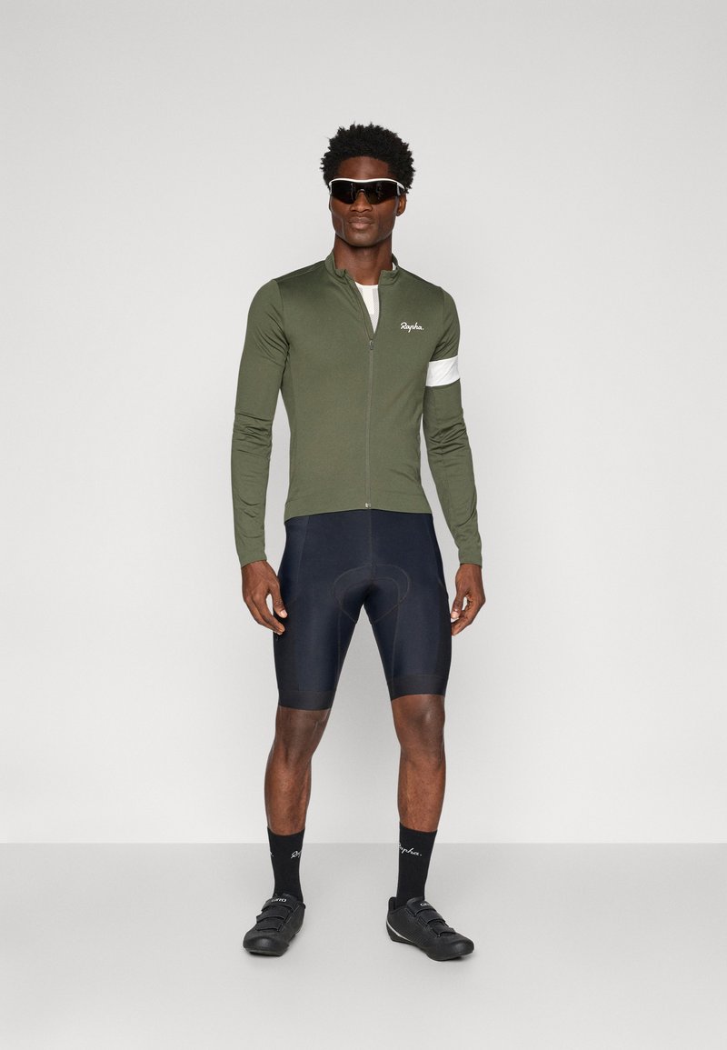 Grön långärmad cykeltröja med dragkedja, vit logotyp och detaljer, ihop med svarta shorts och cykelskor, på en stående figur.