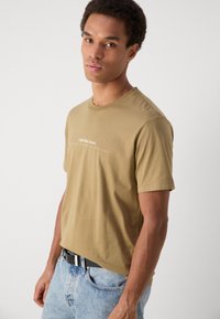 Tan cotton t-shirt with short sleeves and a crew neckline. Features a subtle logo in white across the chest. Worn with light blue jeans.