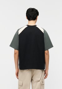 Person med kort svart hår som har på seg en svart og grå kortermet skjorte med blokkmønster og beige cargobukser, sett bakfra.