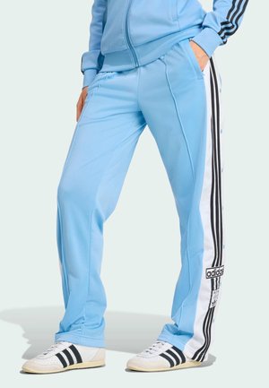 Osoba nosząca jasnoniebieskie spodnie dresowe Adidas z białymi i czarnymi paskami oraz białe buty sportowe Adidas z czarnymi paskami.