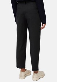 Persoon in zwarte rechttoe-rechtaan broek, marineblauwe trui en beige schoenen met dikke zool, van achteren weergegeven tegen een effen achtergrond.