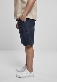 Shorts cargo navy avec plusieurs poches, en tissu texturé. Portés avec des baskets blanches. Le modèle se tient avec une main dans la poche.