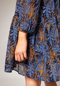Robe à motifs floraux bleus et orange avec tissu transparent ; manches bouffantes et taille froncée. Main visible tenant le tissu.