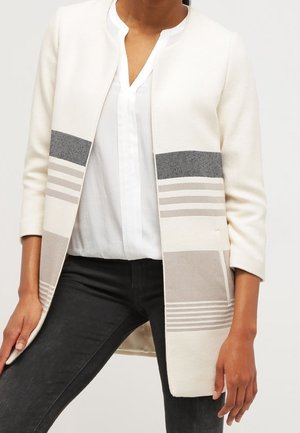 Vrouw draagt een witte openvallende cardigan met grijze strepen, een witte blouse en een zwarte broek, staand tegen een effen achtergrond.