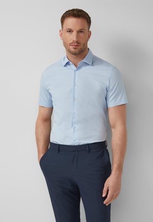 Man met lichtblauw overhemd met korte mouwen en donkere blauwe broek, staand met één hand in de zak tegen een witte achtergrond.