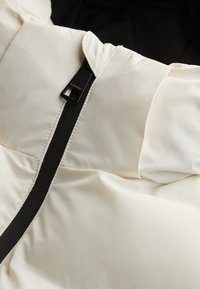 Primer plano de una chaqueta acolchada de color crema con una cremallera vertical negra y una lengüeta con la etiqueta "Superdry" sobre un fondo oscuro.