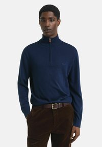 Jonge man in een marineblauwe quarter-zip trui, gestoken in een bruine corduroybroek met een bruine leren riem, staande tegen een effen achtergrond.