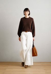 Καφέ πουλόβερ πλεκτό, λευκές παντελόνες με φαρδιά εφαρμογή, καφέ ζώνη, κρατώντας μια καφέ τσάντα tote. Κοντά μαλλιά, απλά σκουλαρίκια, στέκεται σε ξύλινο πάτωμα.