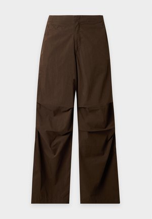 Jeune homme aux cheveux noirs portant un t-shirt noir, un pantalon large marron, des baskets blanches et un sac à bandoulière noir.