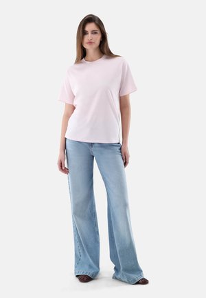 Junge Frau, die ein hellrosa T-Shirt mit kurzen Ärmeln und weit geschnittene, hellblaue Jeans trägt, steht vor einem einfarbigen Hintergrund.