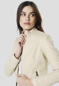 Crèmekleurig leren jack met een aansluitend ontwerp, hoge kraag en textuurelementen op de schouders. Voorzien van een ritssluiting aan de voorkant en zijzakken.