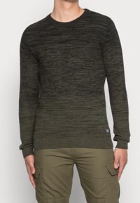 Pull en maille vert foncé avec un col rond, un motif texturé et des poignets et ourlet en côtes. Porté sur un pantalon cargo beige.