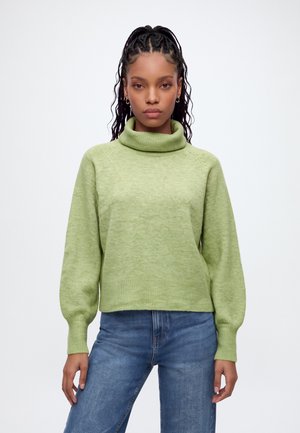 Femme aux cheveux tressés portant un pull à col roulé vert et un jean bleu, debout sur fond blanc.