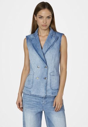 Frau trägt eine hellblaue ärmellose, doppelreihige Jeansweste und passende Jeans, steht vor einem schlichten weißen Hintergrund.