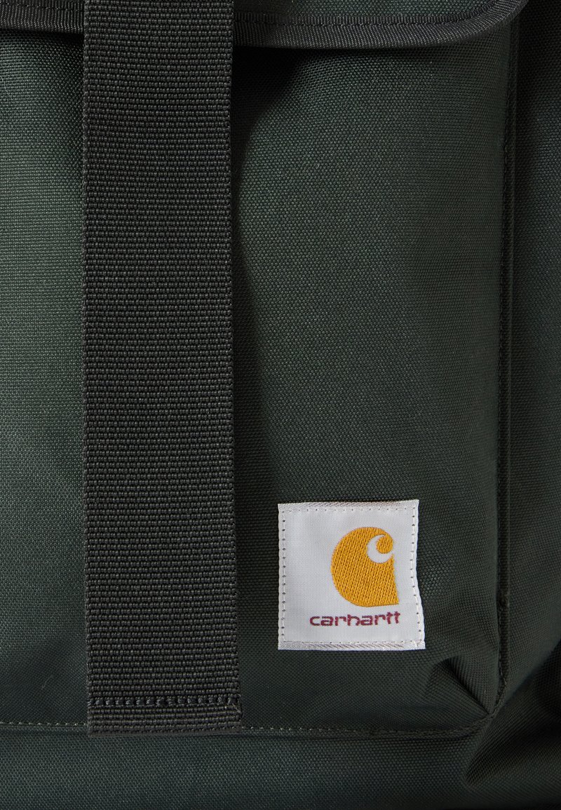 Dunkelgrüner Stoff mit schwarzem Riemen und weißem Carhartt-Logo-Patch mit gelbem Symbol und rotem Text auf einer Tasche.