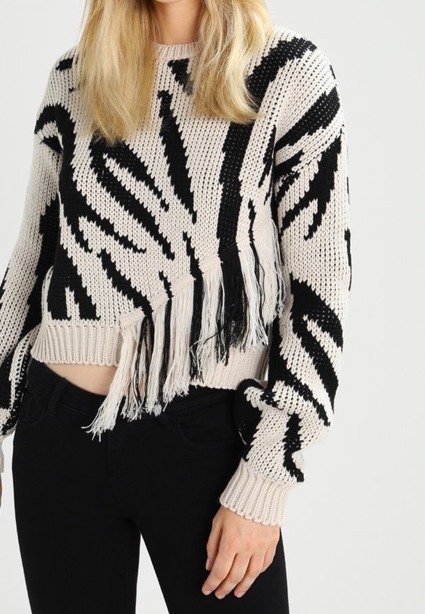 Krátký pletený svetr v béžové barvě s černými zigzagovými vzory. Obsahuje třásně na spodním okraji a žebrované manžety. Povrch vypadá měkce.