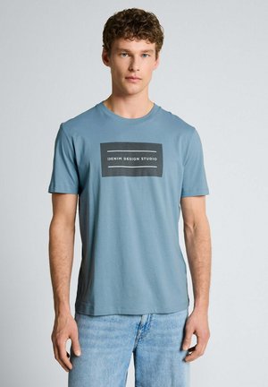 Junger Mann mit lockigem Haar, der ein blaues "Denim Design Studio" T-Shirt und hellblaue Jeans trägt und vor einem schlichten weißen Hintergrund steht.