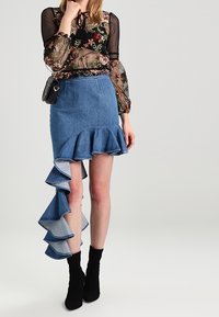 Gonna di jeans con orlo a ruffle ondulato, colore blu scuro, abbinata a una blusa floreale trasparente e stivali neri alla caviglia. Borsa a tracolla visibile.