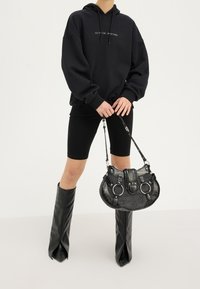 Sweat à capuche noir oversize avec texte, associé à un cuissard noir et des bottes noires brillantes montant jusqu'aux genoux. Tenant un sac à main noir texturé avec des accents.