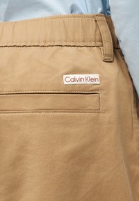 Κοντινό πλάνο στην τσέπη μπεζ παντελονιού με ετικέτα Calvin Klein ραμμένη πάνω από την τσέπη σχισμής, φορεμένο πάνω από ένα ανοιχτό μπλε πουκάμισο.