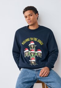 Marinblå sweatshirt med en grafik som visar dödskallar med tomteluvor mot ett grönt kors, texten lyder "WELCOME TO THE JINGLE," ribbade muddar.