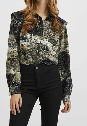 Langärmlige Bluse mit einem schwarz-grünen abstrakten Muster, Knopfverschluss und rüschendetails an den Schultern, kombiniert mit schwarzen hochgeschnittenen Hosen.