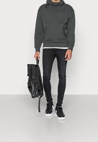 Felpa con cappuccio grigio scuro con tasca frontale, abbinata a jeans skinny neri e scarpe da ginnastica nere. L'individuo tiene uno zaino nero.