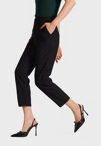 Pantaloni neri su misura con vestibilità slim, dotati di tasche laterali. Abbinati a décolleté nere con punta appuntita e dettagli a fiocco.