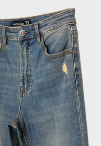 Vaqueros de mezclilla azul claro con un patrón desgastado, que presentan un área descolorida y herrajes en tono cobre. Diseño estándar de cinco bolsillos.