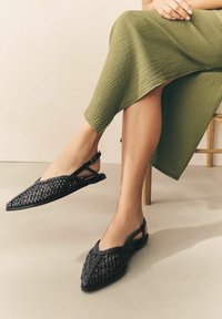 Juodos audinės slingback bateliai su smailiu nosimi ir plokščiu padu, turintys reguliuojamus dirželius ir tekstūruotą paviršių.